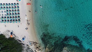 Luchtbeeld van een strand met helder turquoise water, een rij kleurrijke parasols en mensen die van het strand genieten.