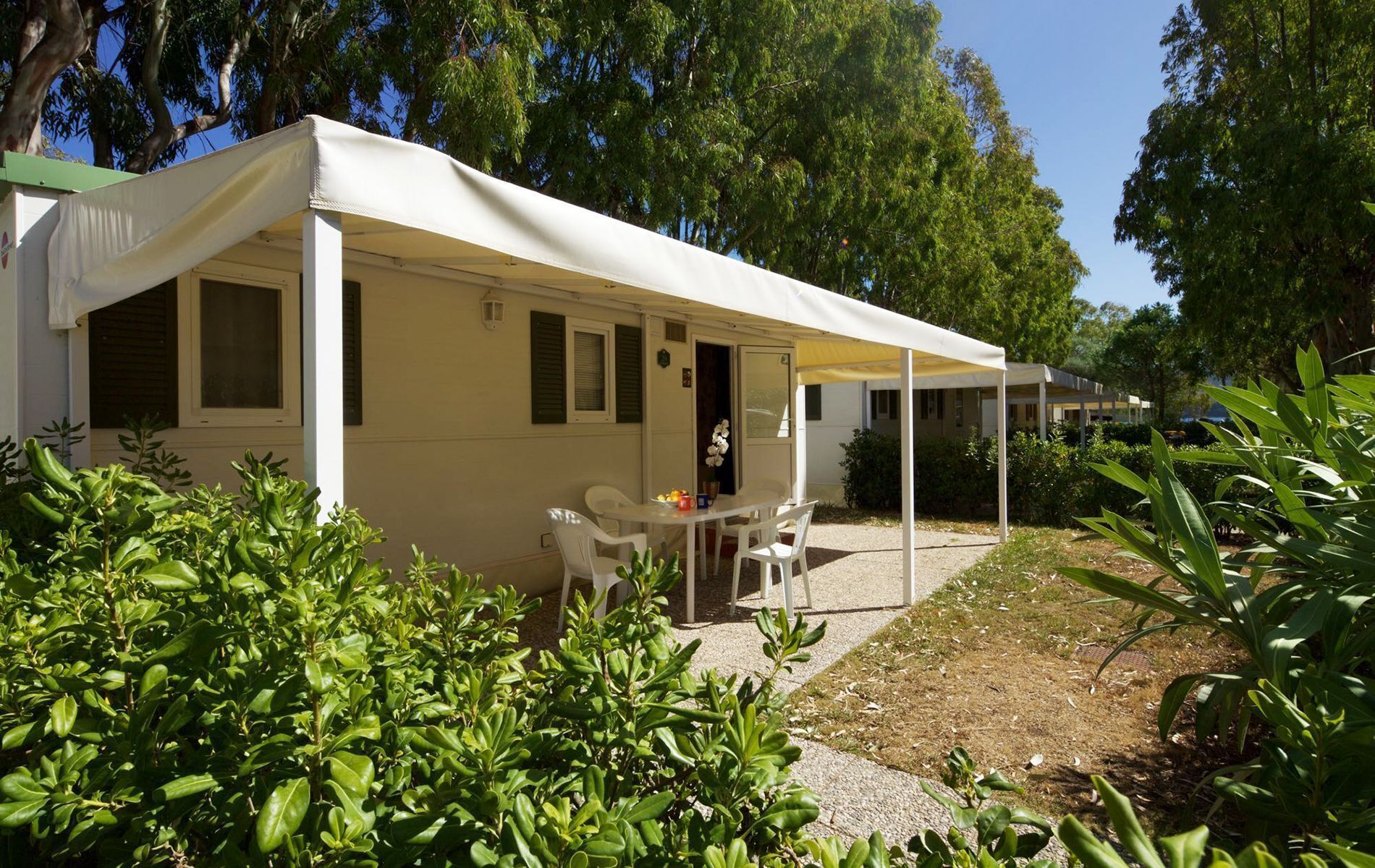 A single-story mobile home with a covered patio area, a dining table with chairs, and a small garden area with various plants.