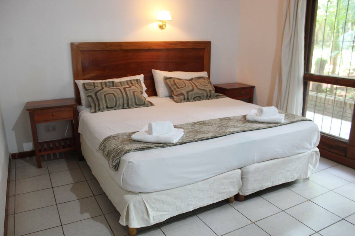 A neatly made bed with white linens, two pillows, and a blanket. There is a nightstand with a lamp on it. The room has a tiled floor and a window with curtains.