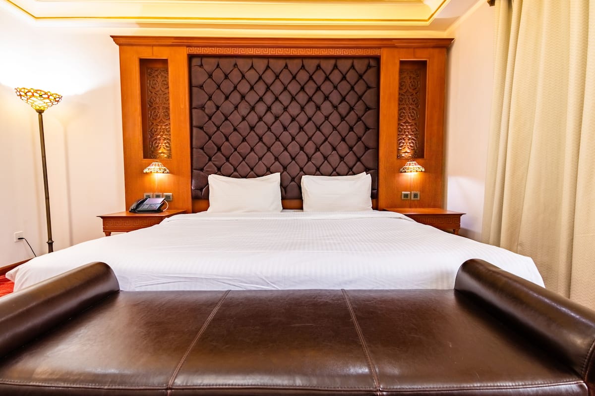 A hotel room with a large bed, a headboard with a diamond pattern, two bedside lamps, and a brown leather ottoman.