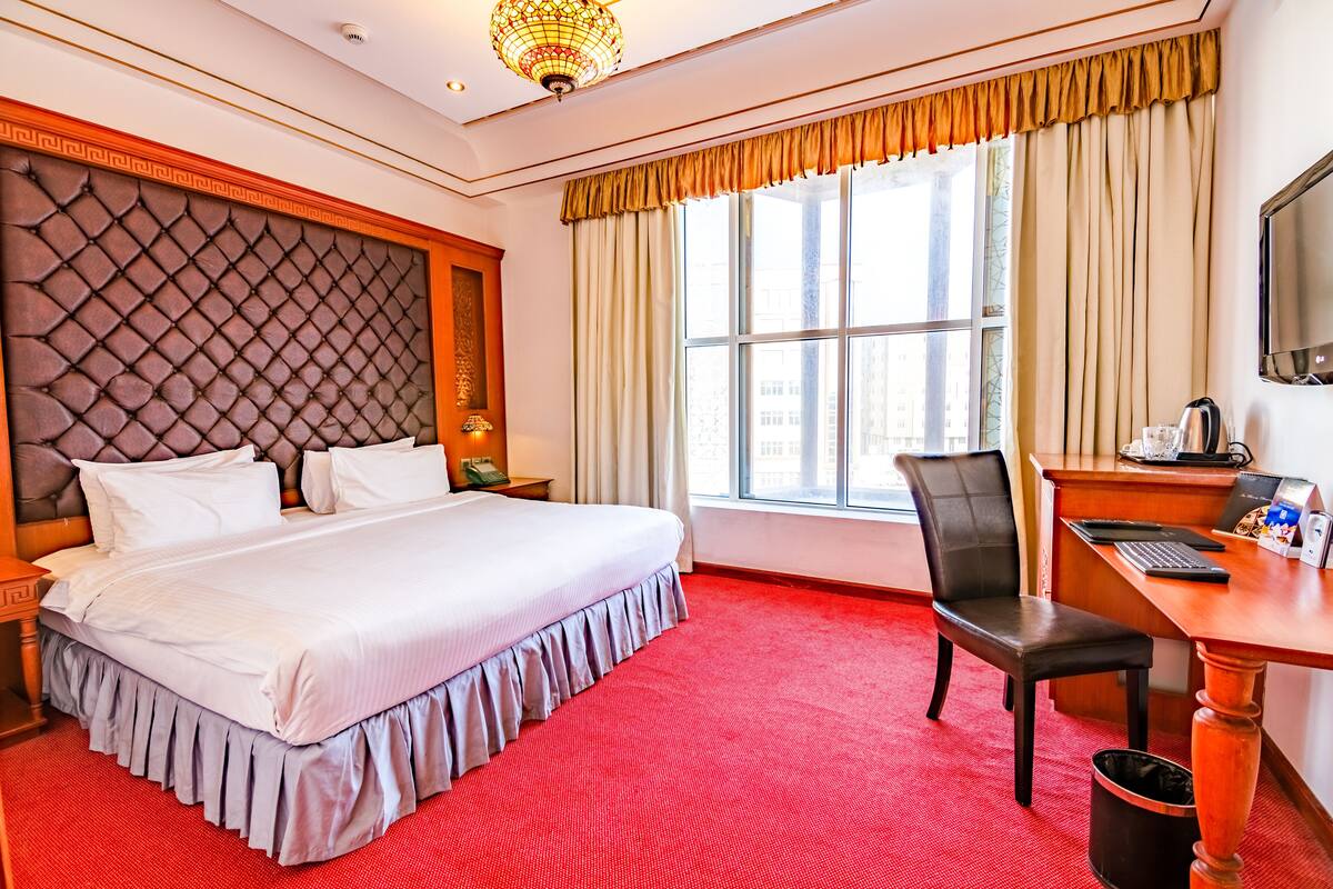 A hotel room with a large bed, a desk with a computer, a chair, and a window with curtains.