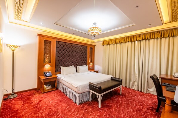 A hotel room with a large bed, a desk with a chair, a red carpeted floor, a chandelier, and a curtain with a golden trim. - Platinum Hotel (Muscat)