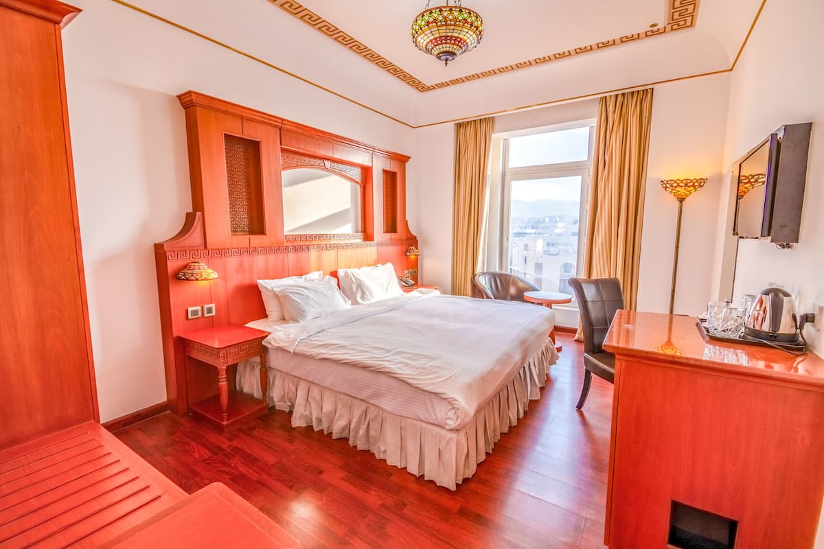 A hotel room with a large bed, a wooden headboard, a desk with a lamp, and a view of the city.