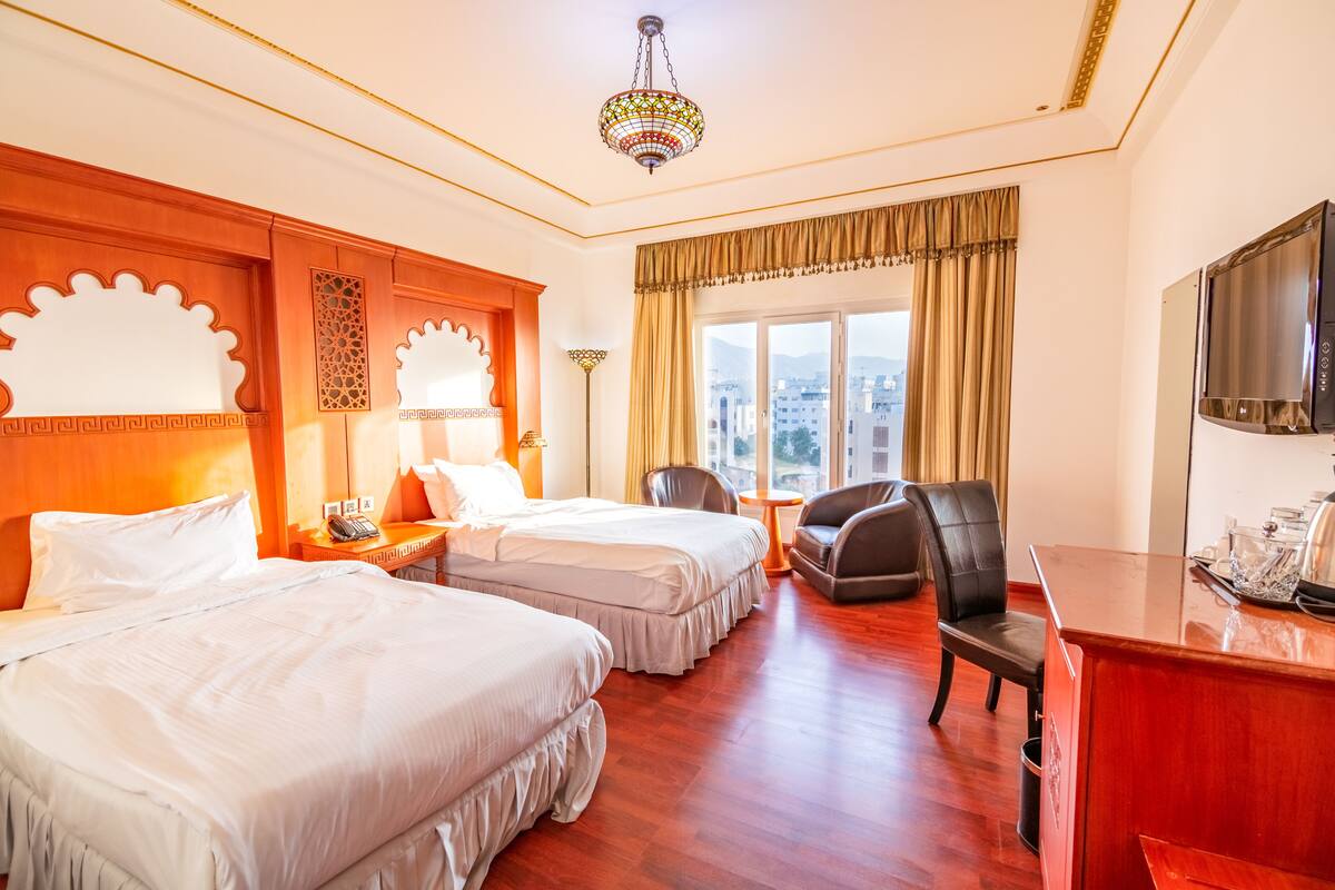 A hotel room with two beds, a desk, a chair, a TV, and a view of the city.