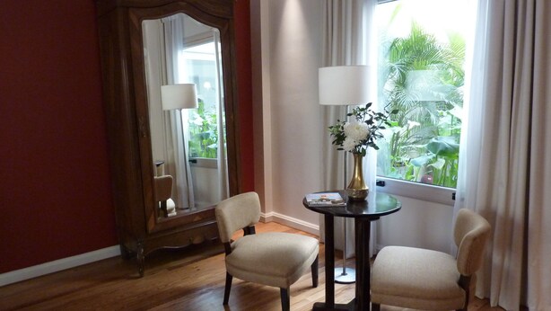 A room with wooden flooring, a large mirror, two chairs, a small round table with a lamp, and a window with curtains. - Magnolia Hotel Boutique (Buenos Aires)