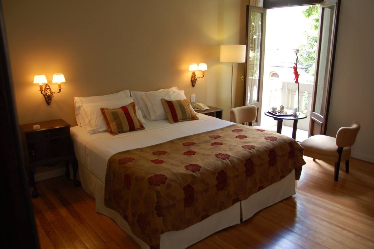 A hotel room with a large bed, bedside tables, a chair, a small table with a lamp, and a view of the outdoors.