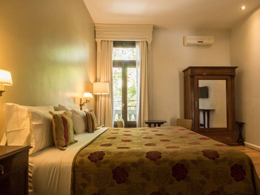 A hotel room with a bed, two bedside lamps, a large mirror, a small table, and a view of the outdoors through a window.