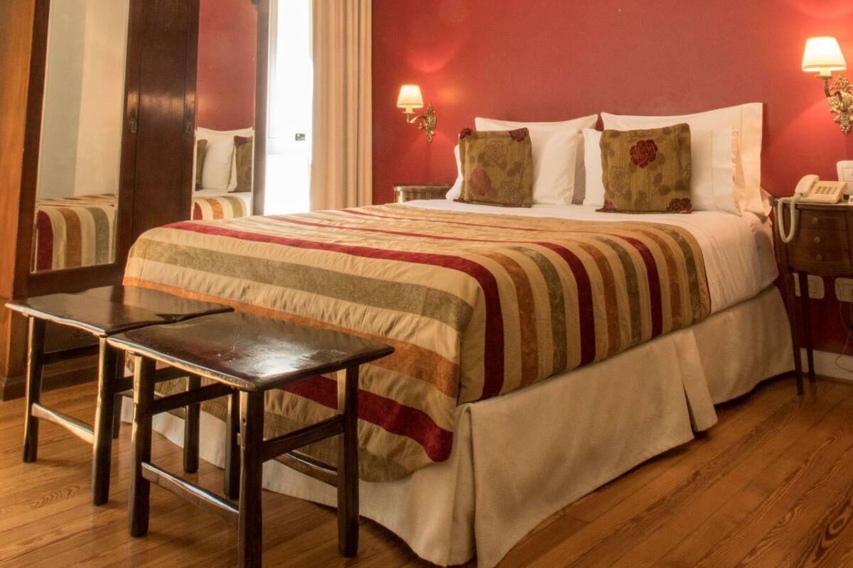 A hotel room with a wooden bed, a desk, a chair, a mirror, and a red wall.