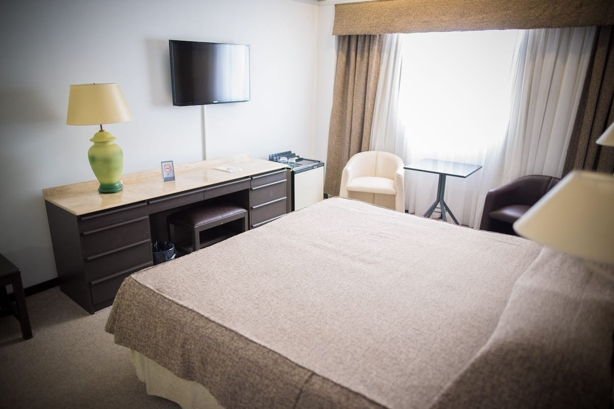 A hotel room with a bed, a desk with a lamp, a chair, a TV on the wall, and a window with curtains.