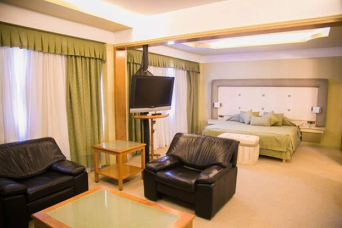 A hotel room with a bed, a television, two leather chairs, a small table, and a glass coffee table.