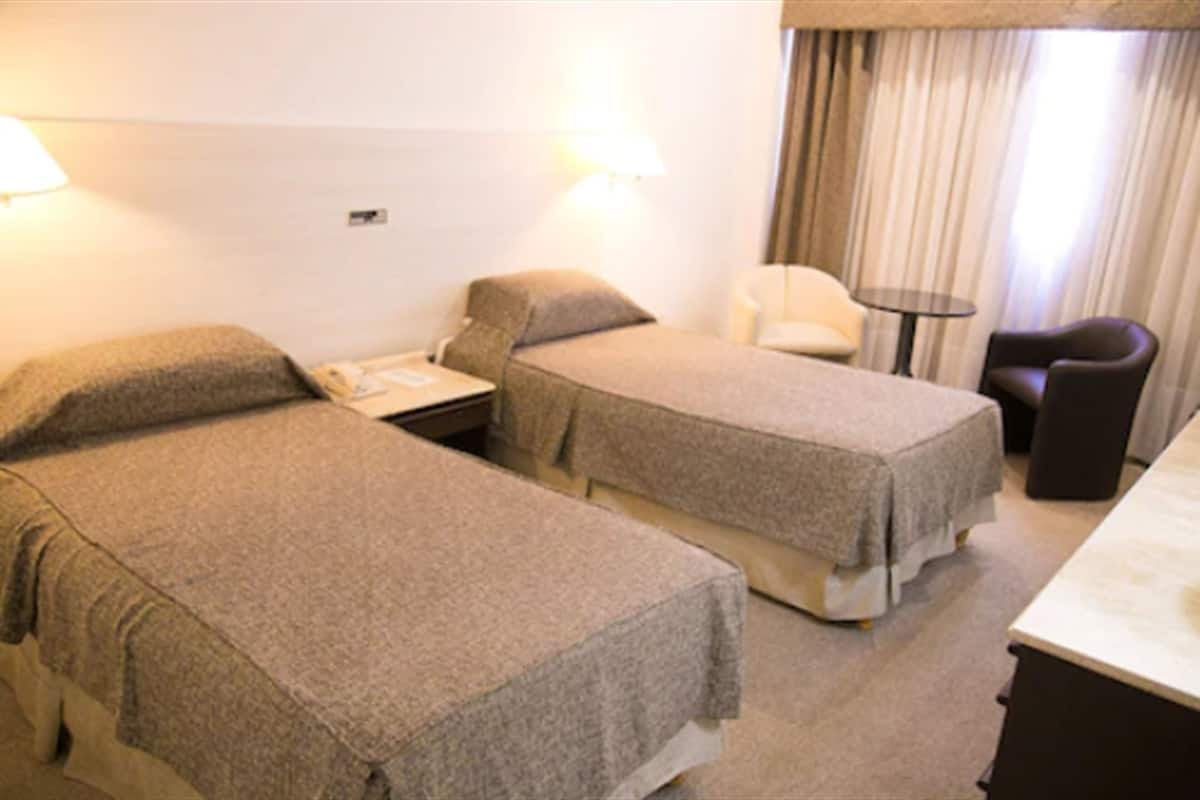 A hotel room with two single beds, a small table, a chair, and a lamp.