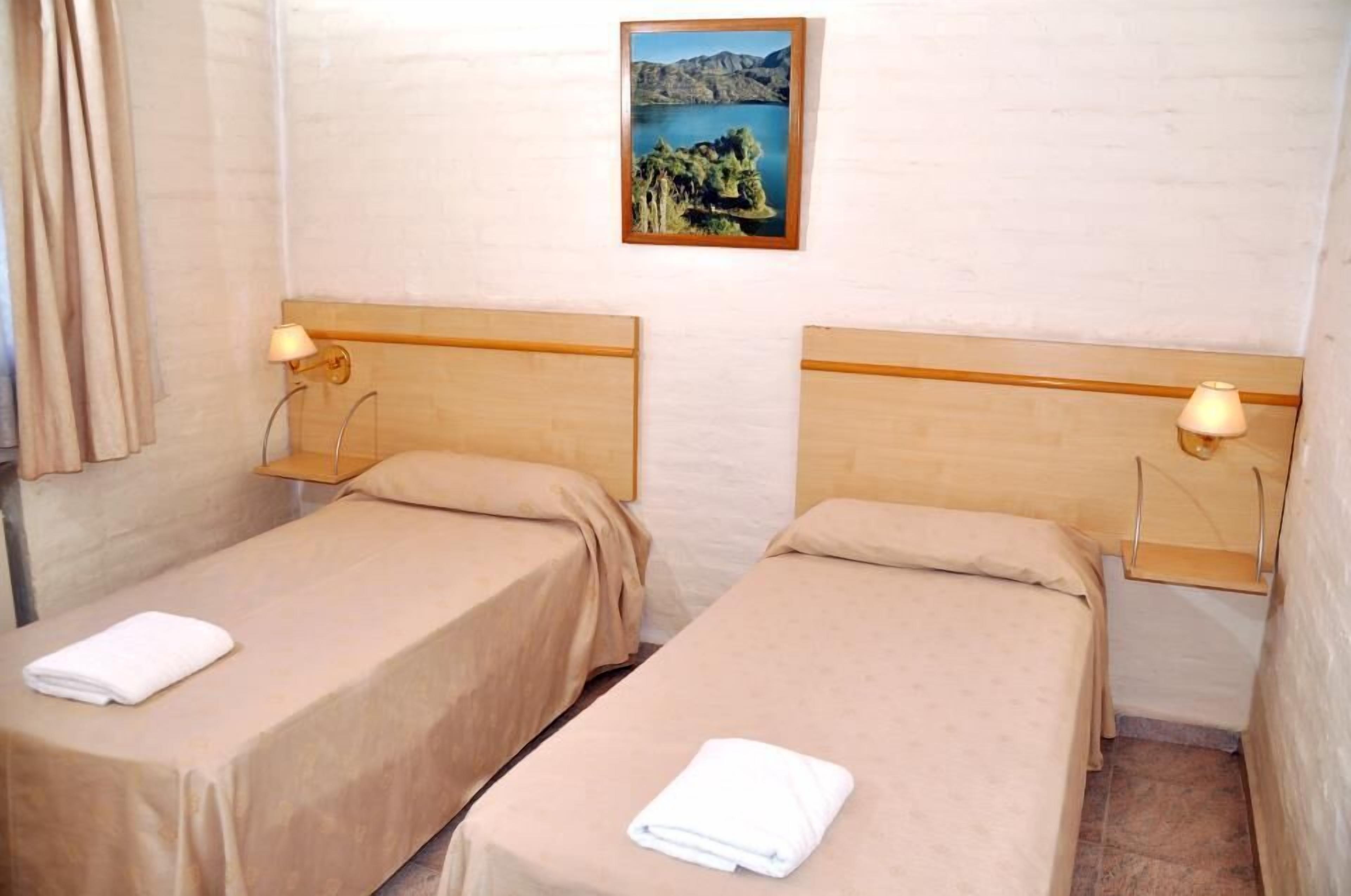 A hotel room with two single beds, each with a wooden headboard and a small bedside table with a lamp. A framed picture of a lake and trees hangs on the wall above the beds.