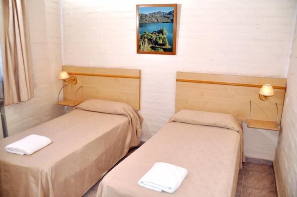 A hotel room with two single beds, each with a wooden headboard and a small bedside table with a lamp. A framed picture of a lake and trees hangs on the wall above the beds.