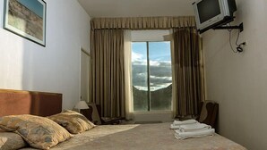 A hotel room with a bed, a television mounted on the wall, a window with curtains, and a mountain view. - Hostería y Cabañas Valle Fertil (San Agustin de Valle Fertil)