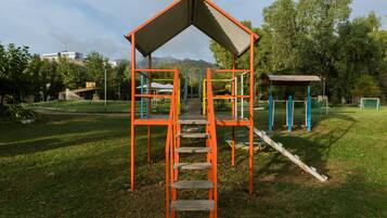 Children can climb to the top of this playground structure, surrounded by a lush green field and shaded by trees.