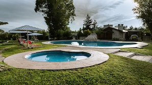A backyard with two swimming pools, a stone house, and a covered seating area with red chairs. - Hostería y Cabañas Valle Fertil (San Agustin de Valle Fertil)
