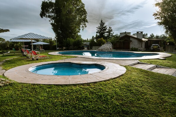 A backyard with two swimming pools, a stone house, and a covered seating area with red chairs.