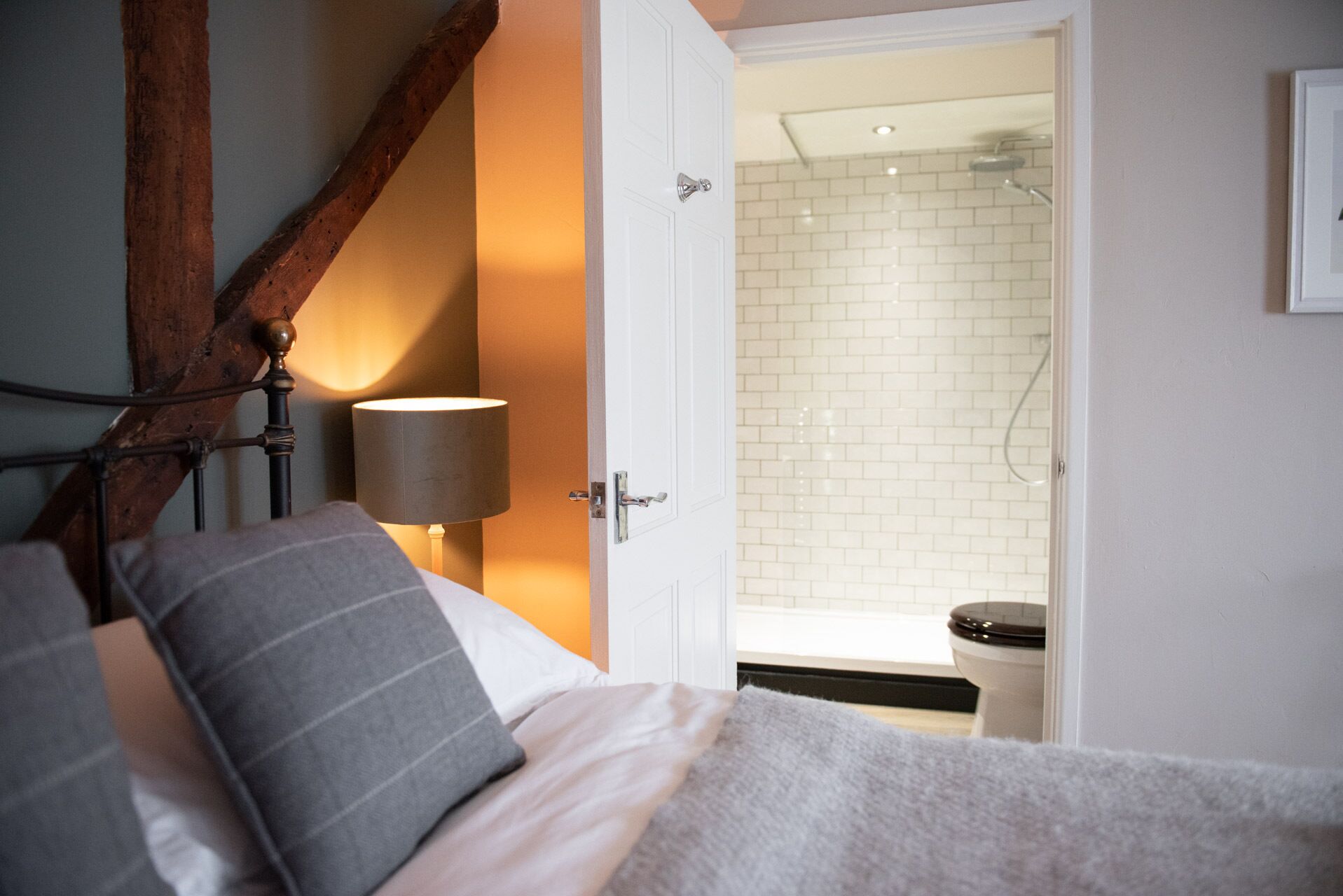 Une chambre à coucher avec un lit, une lampe de chevet, une poutre en bois et une vue sur une salle de bain équipée d’une douche et de toilettes.