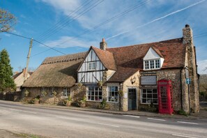 A traditional stone building with a red telephone booth, a sign for 'The Talkhouse' offering dining and bedrooms, and a thatched-roof house. - The Talkhouse (Oxford)