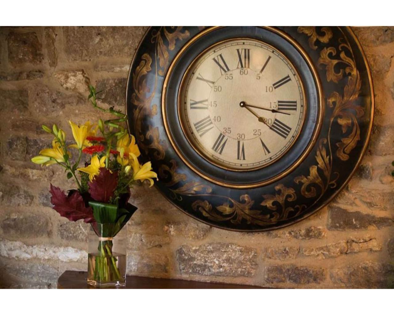 A wall-mounted clock with Roman numerals and a decorative frame, next to a vase with flowers.