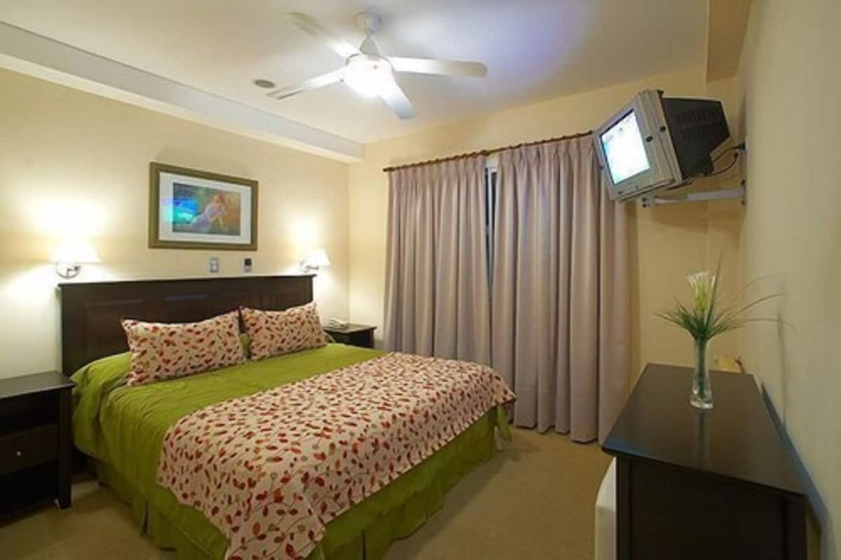 Quarto de hotel com cama, ventoinha no teto, televisão em suporte, mesa de cabeceira e janela com cortinas.