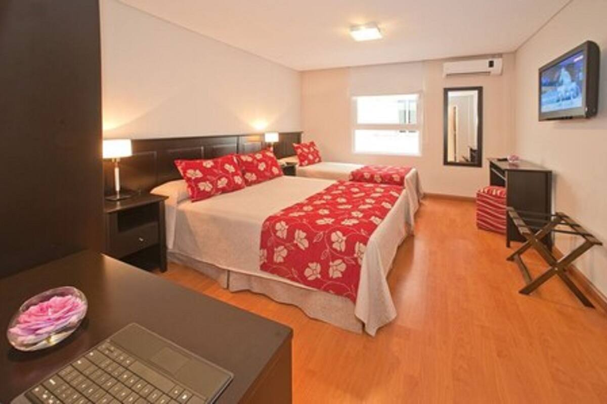 Quarto de hotel com duas camas, uma escrivaninha com um laptop, uma televisão e uma mesinha com uma tigela de frutas.