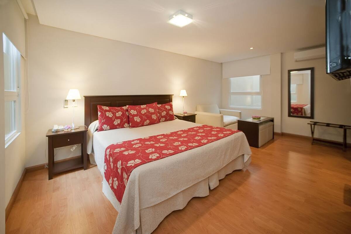 Quarto de hotel com uma cama grande, duas mesas de cabeceira com luminárias, uma cadeira, uma escrivaninha e uma televisão na parede.