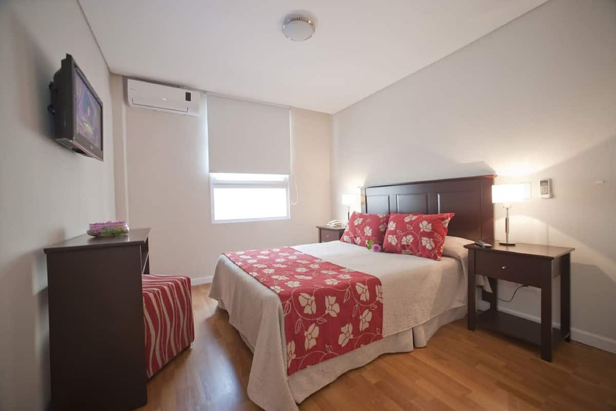Quarto com cama, mesas de cabeceira, criado-mudo, janela com persianas e televisão na parede.
