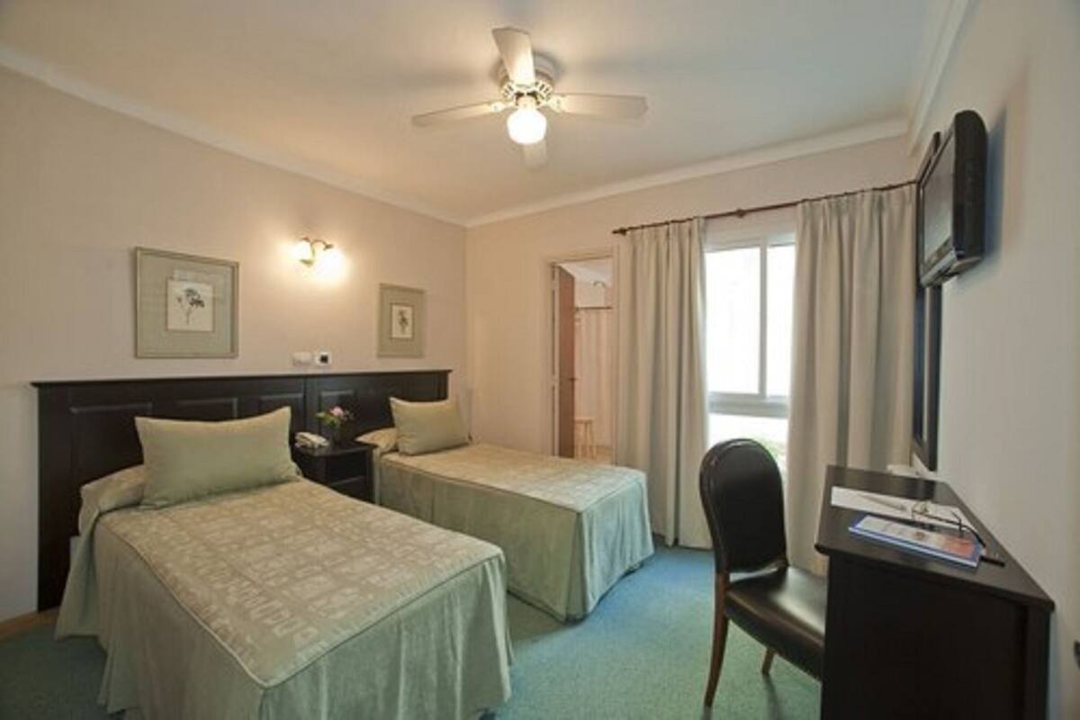 Quarto de hotel com duas camas de solteiro, uma escrivaninha com cadeira, ventoinha no teto e janela com cortinas.
