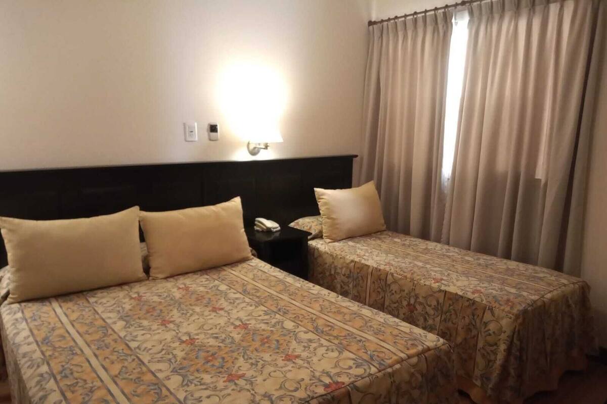 Quarto de hotel com duas camas, uma mesa de cabeceira, uma luminária e uma janela com cortinas.