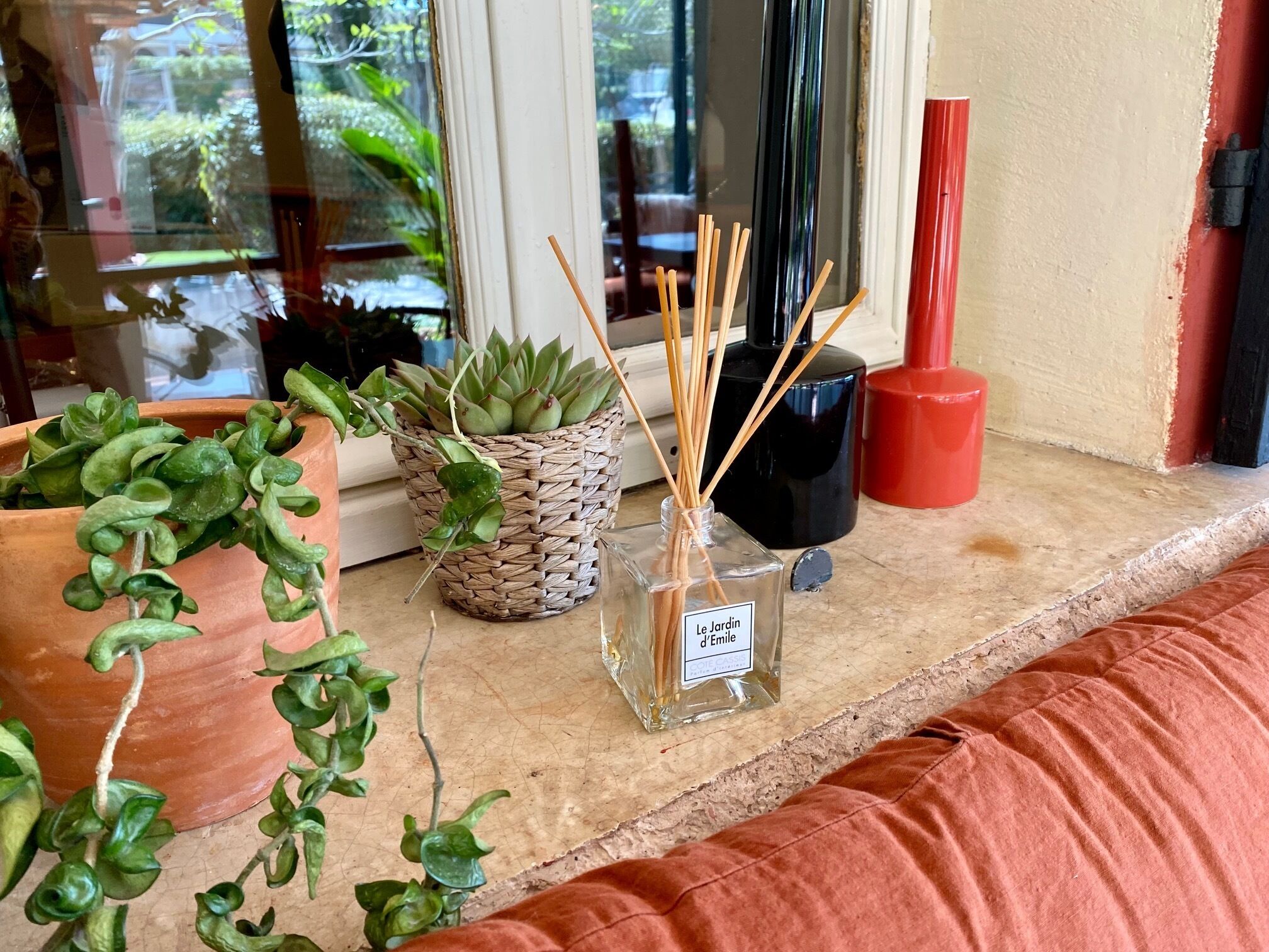 A window sill with potted plants, a reed diffuser, and a red candle holder.