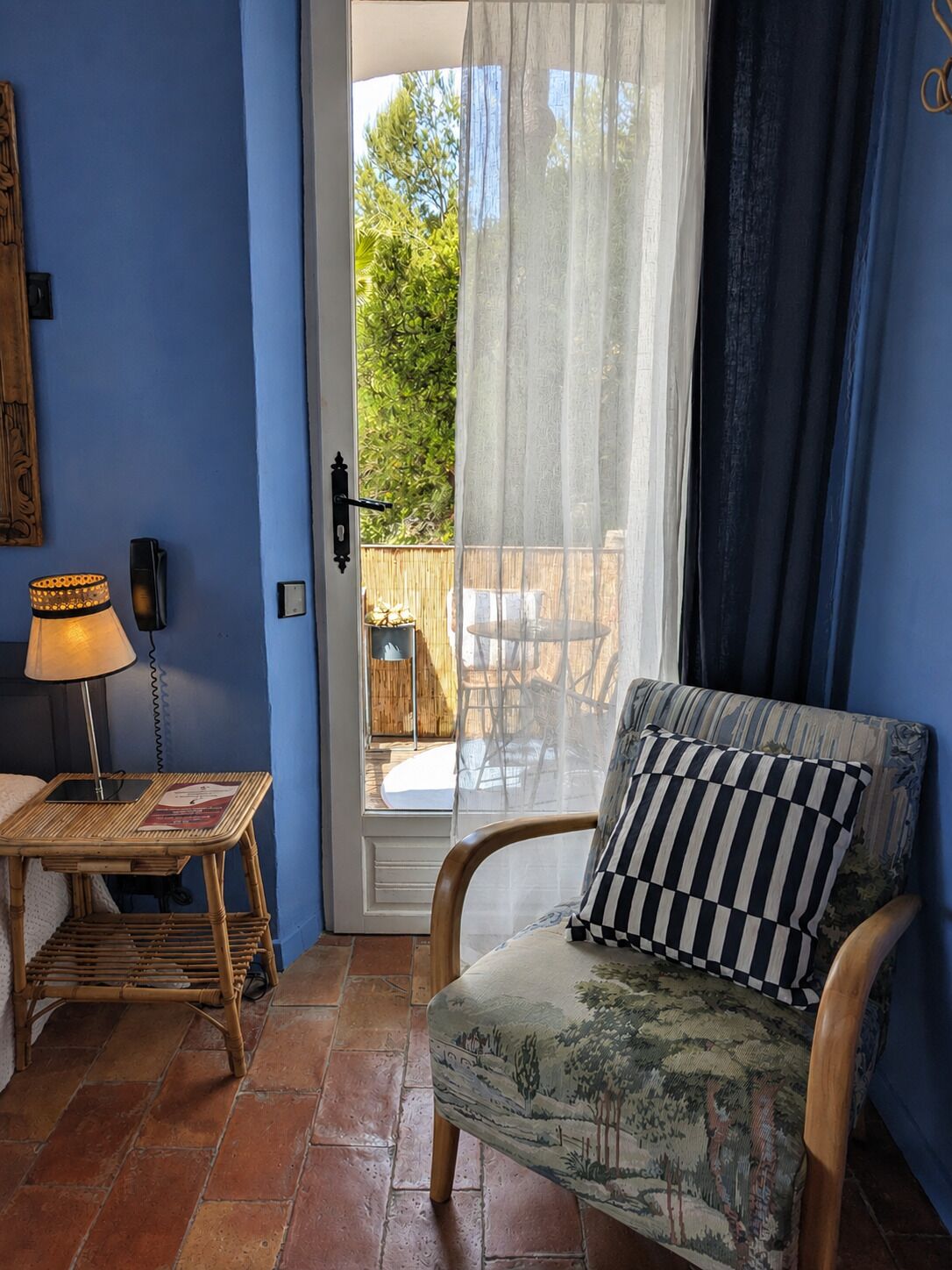 A room with a blue wall, a wicker side table with a lamp, a chair with a checkered pillow, and a view through a glass door to an outdoor area with a table and chairs.