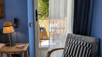 Ein Zimmer mit blauer Wand, einem Korbstuhl mit Lampe, einem Stuhl mit kariertem Kissen und Blick durch eine Glastür auf einen Außenbereich mit Tisch und Stühlen.