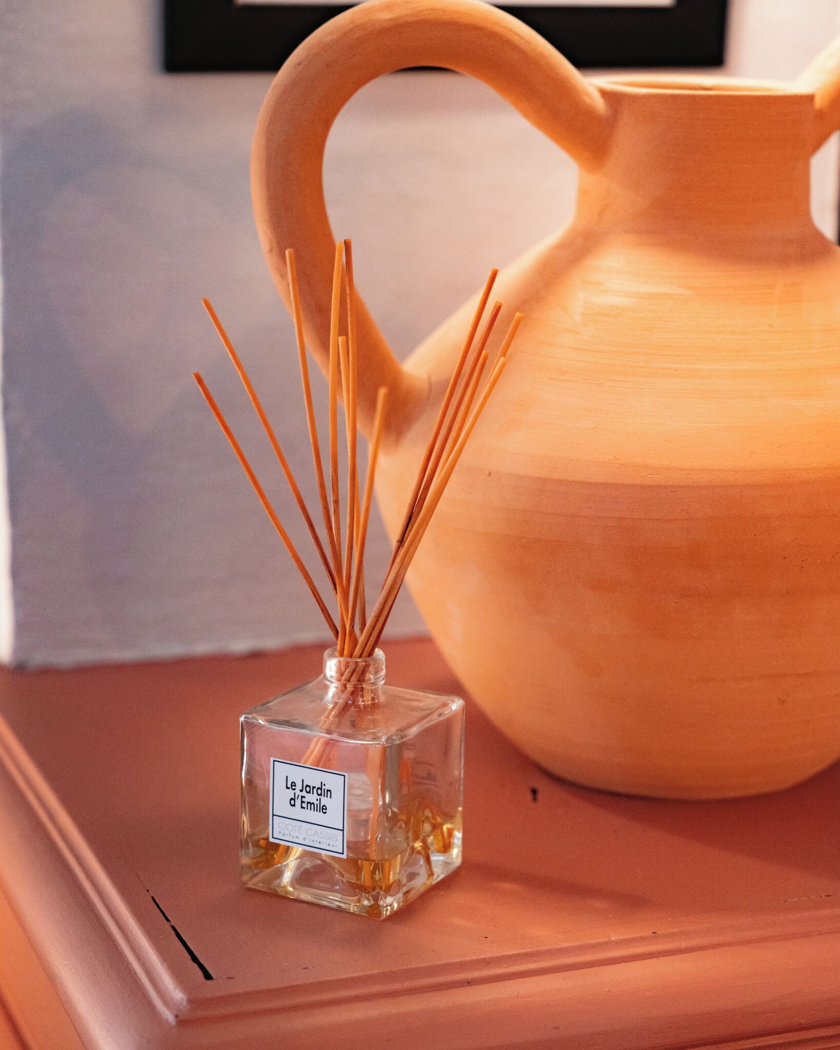 Eine klare Glasflasche mit Schilfrohrstäbchen und einem Etikett, platziert auf einer Holzoberfläche neben einer Keramikkanne.