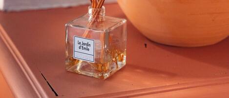 Eine klare Glasflasche mit Schilfrohrstäbchen und einem Etikett, platziert auf einer Holzoberfläche neben einer Keramikkanne.