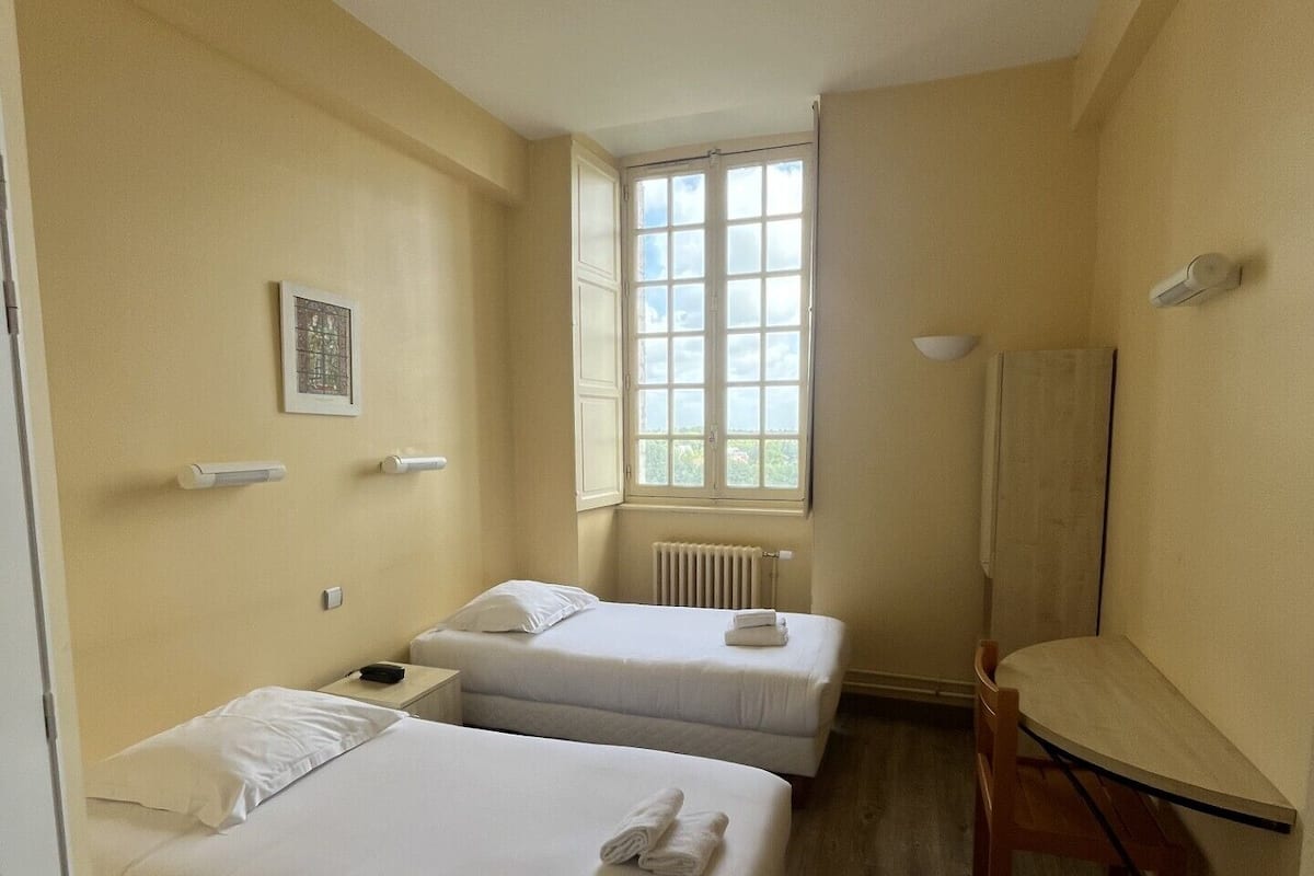 A hotel room with two beds, a radiator, a window, and a desk.