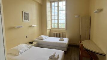 A hotel room with two beds, a radiator, a window, and a desk.