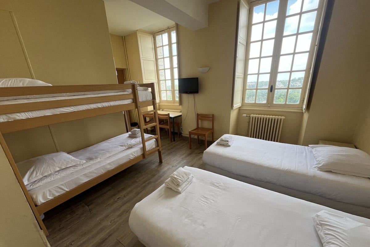 A dormitory room with bunk beds, a desk, a chair, a TV, and a window with a view.
