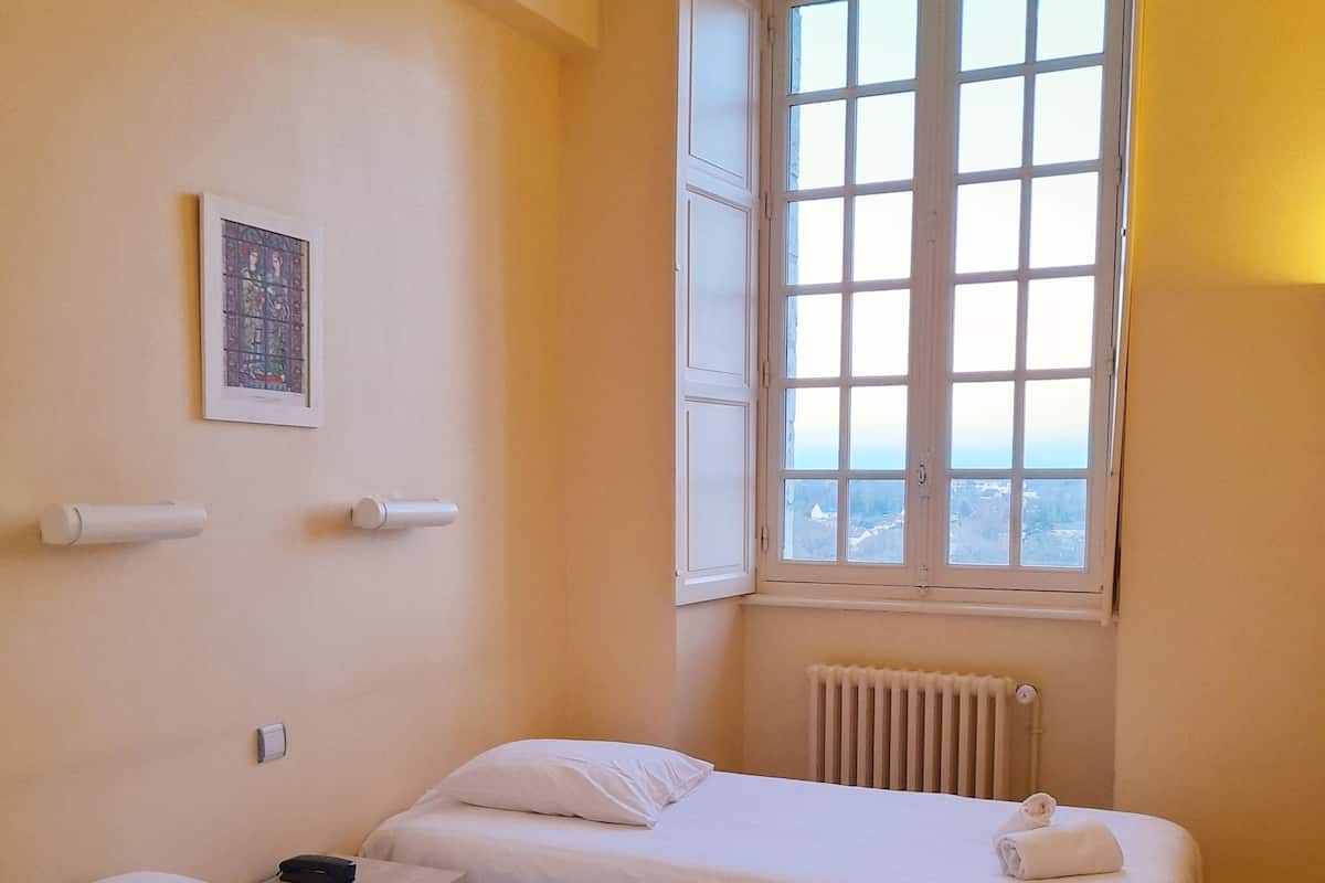 A hotel room with two single beds, a radiator, a window with a view, and a framed picture on the wall.