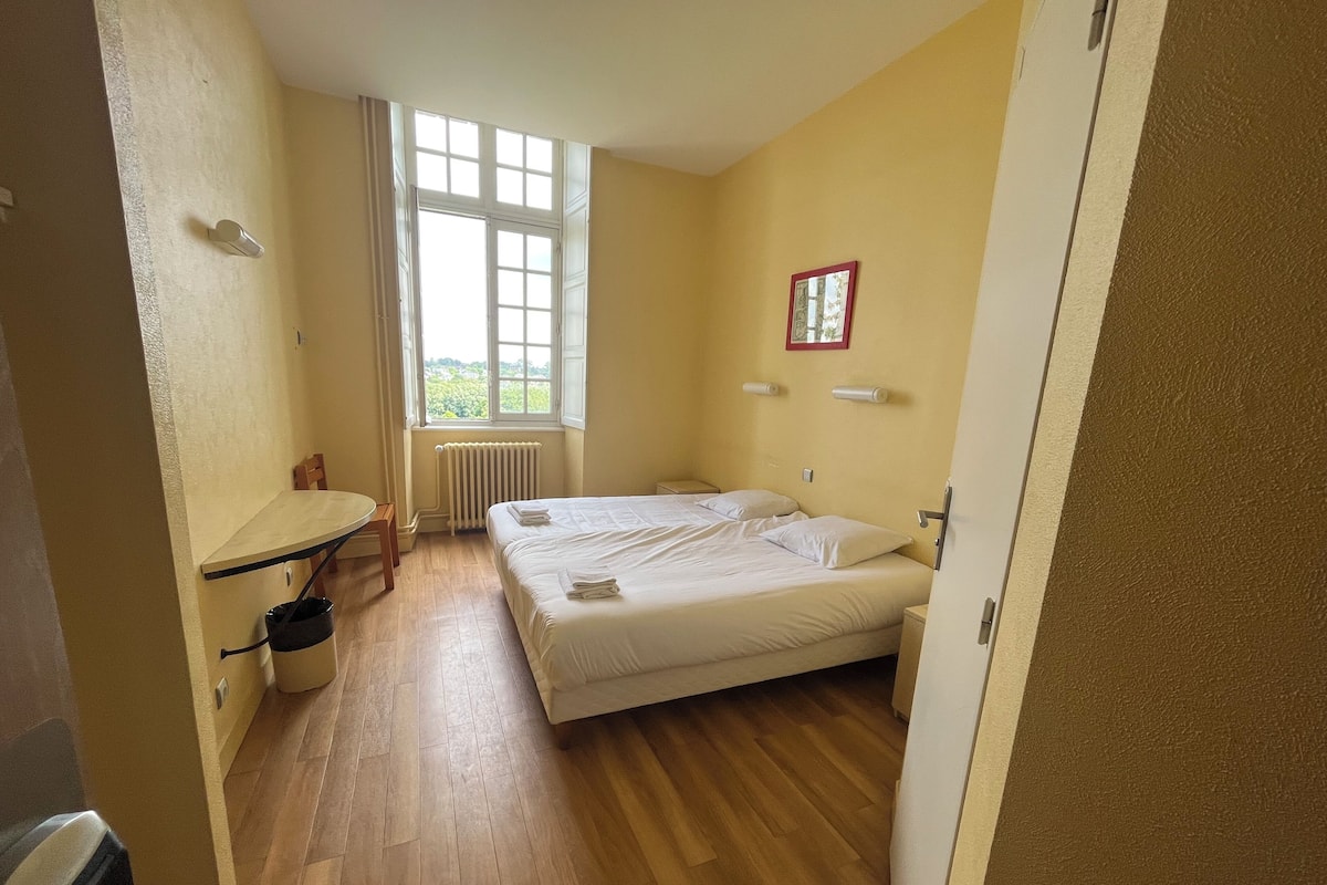 A bedroom with a double bed, a small table, a radiator, and a window with a view.
