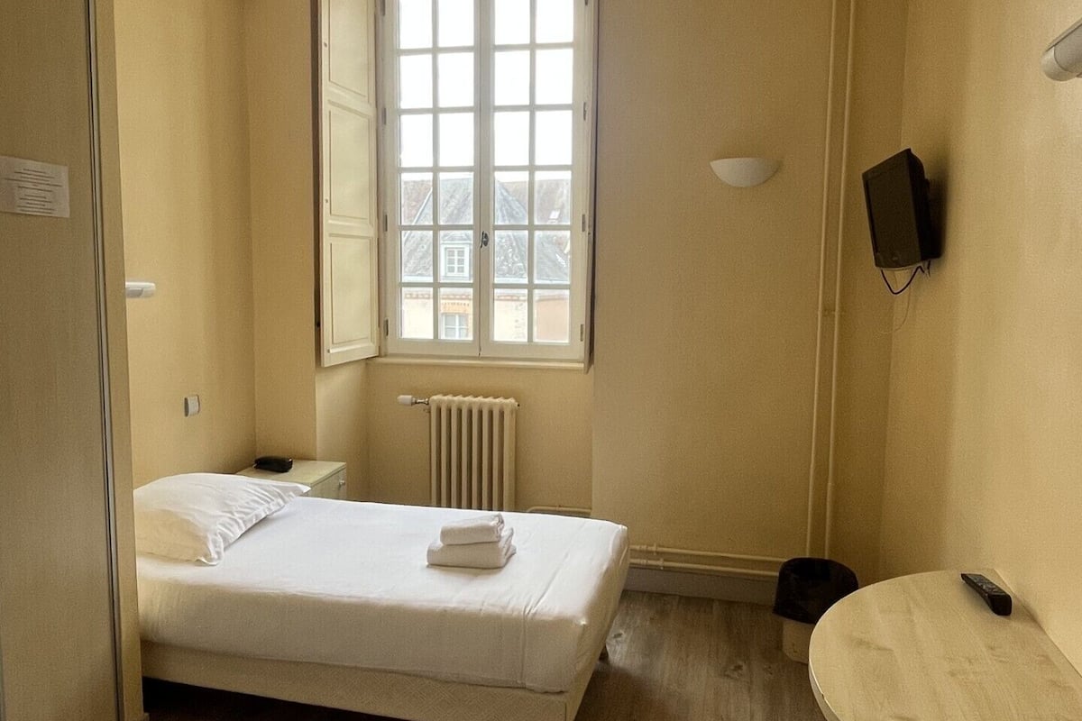 A small, compact room with a single bed, a round table, a radiator, and a window with a view of buildings outside.