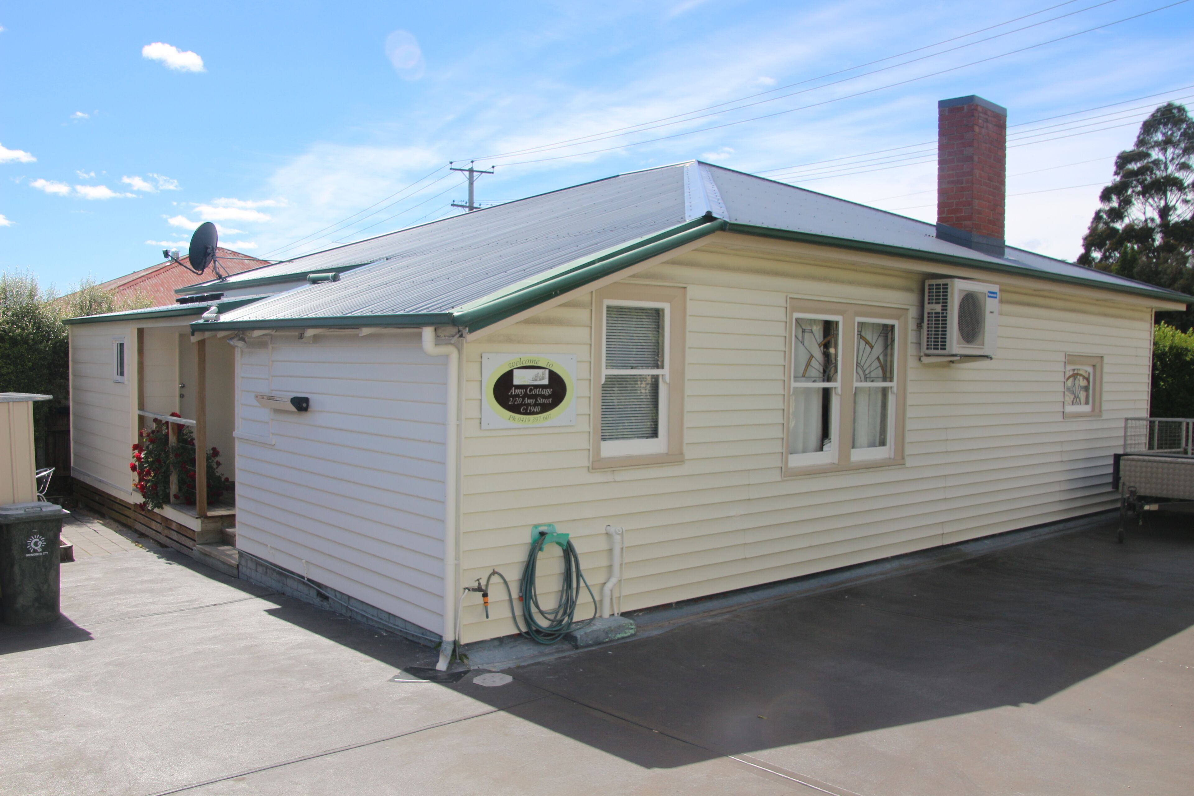 A small, single-story building with a green roof and beige siding, featuring a front door, windows, and a sign that reads "Amy Cottage."