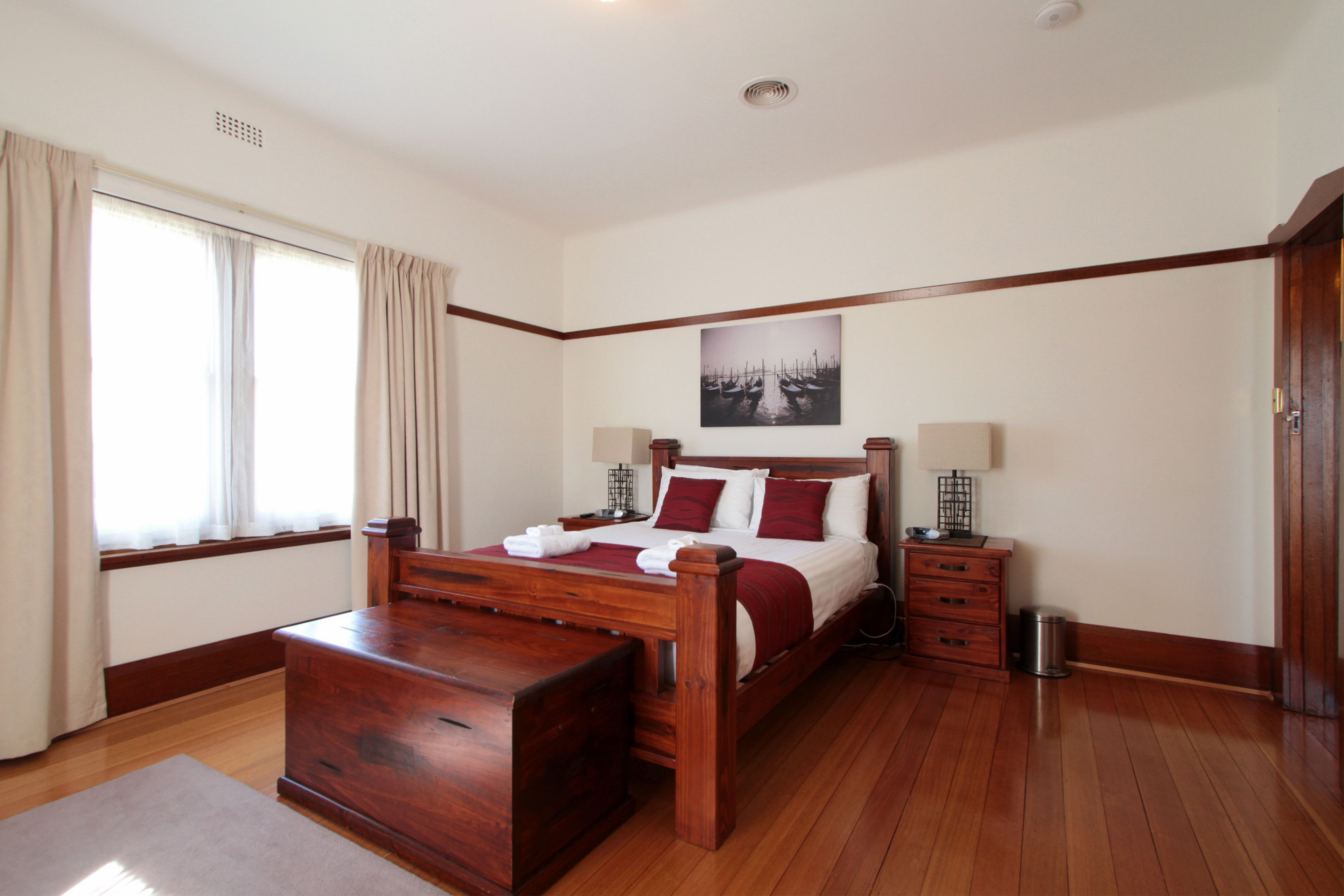 Un dormitorio con una cama de madera, una mesita de noche, una lámpara de noche, una ventana con cortinas y un cuadro colgado en la pared.