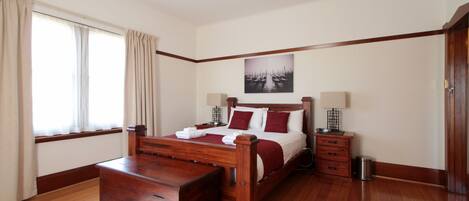 Un dormitorio con una cama de madera, una mesita de noche, una lámpara de noche, una ventana con cortinas y un cuadro colgado en la pared.