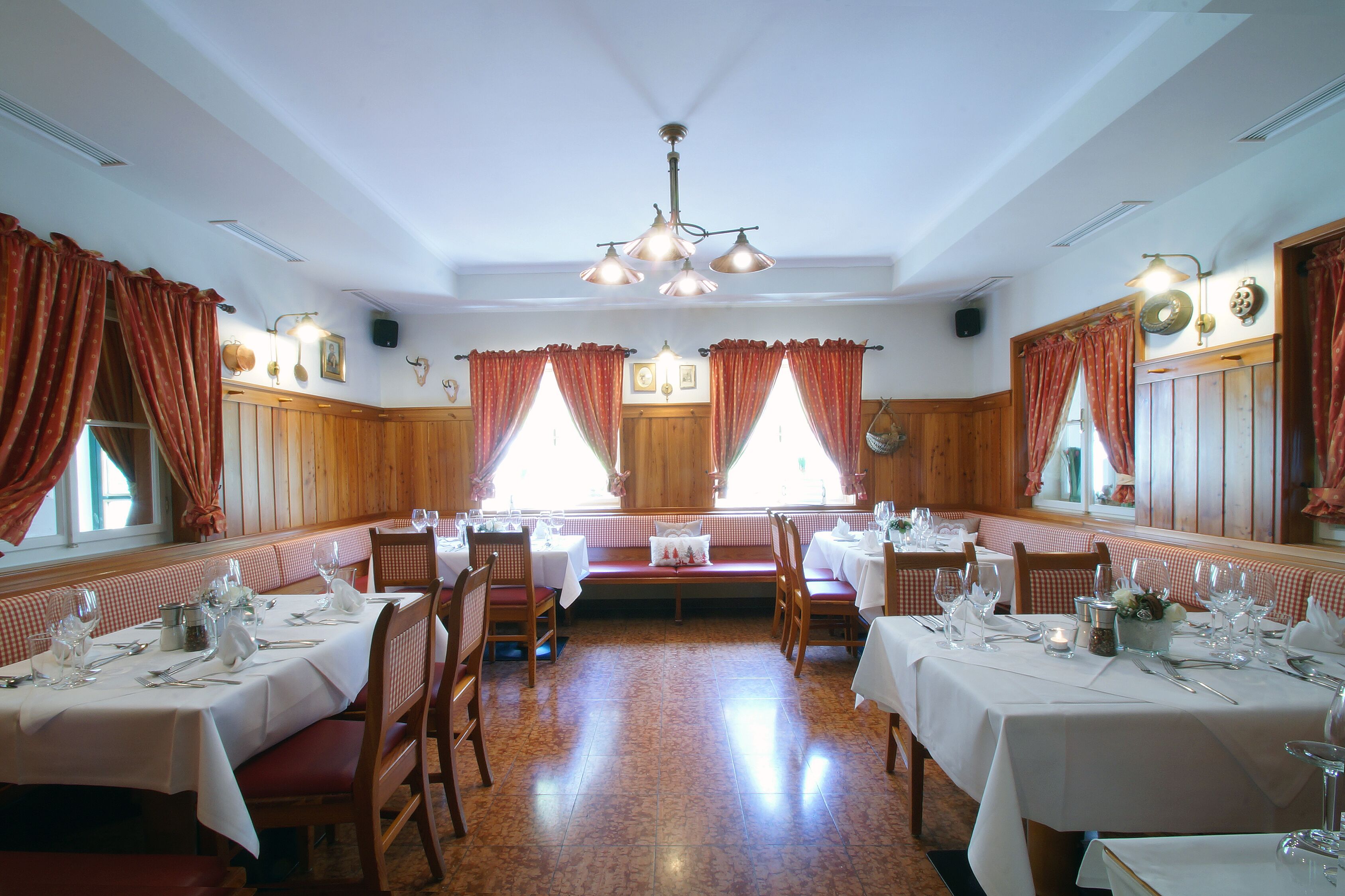 Un ristorante con pannelli in legno, tende rosse e tovaglie bianche. I tavoli sono apparecchiati con bicchieri e piatti.