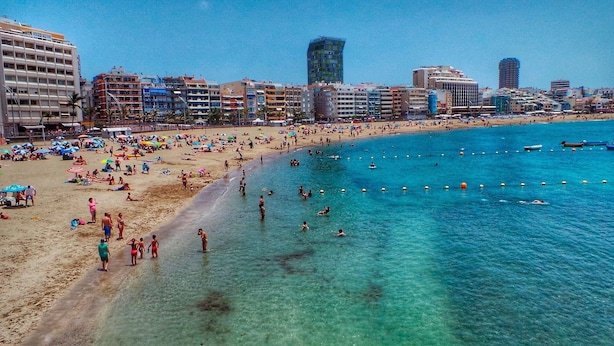 Een strand met helderblauw water, mensen die zwemmen en zonnen, en een stadsgezicht op de achtergrond.