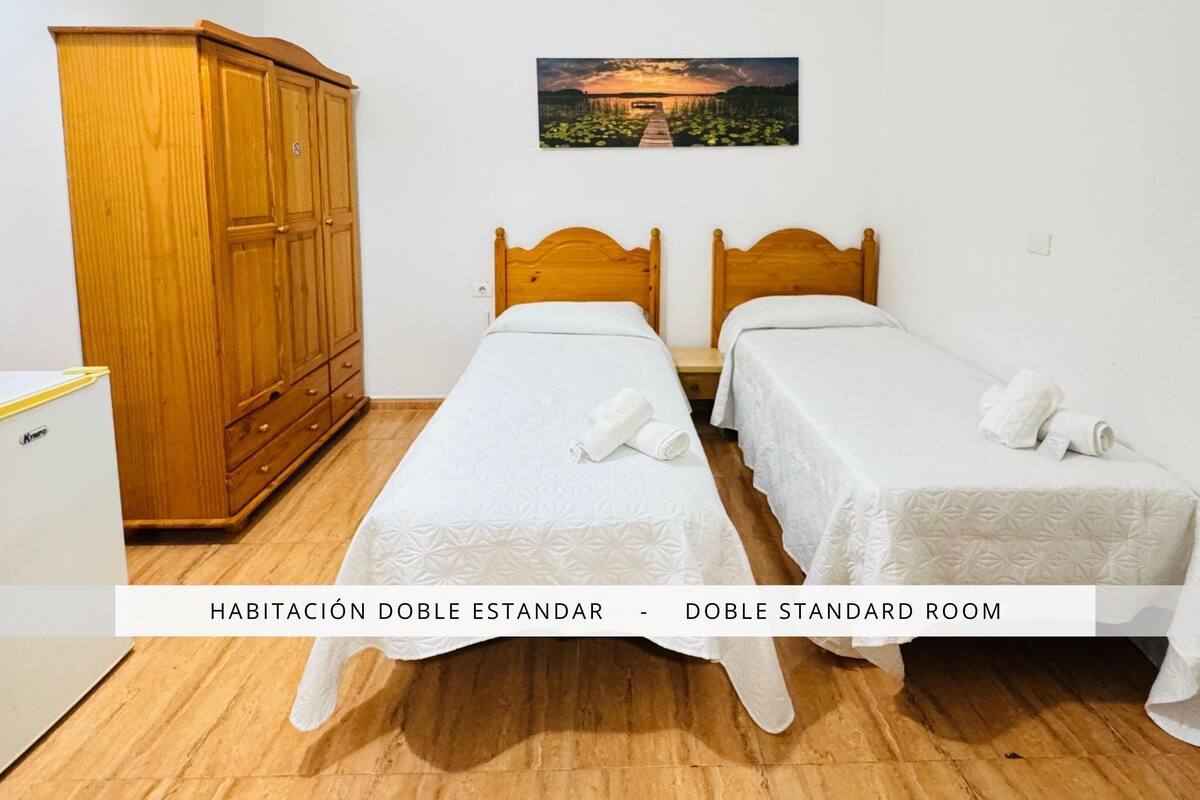 Habitación doble con dos camas individuales, un armario de madera y un refrigerador blanco pequeño.