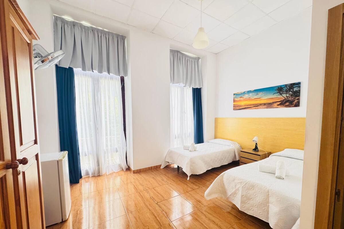 Habitación de hotel con dos camas, piso de madera, ventilador de techo y un cuadro de un atardecer.
