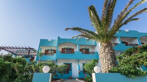 A multi-story building with a blue facade, balconies, and a palm tree in the foreground. - Alpha Studios (Karpathos)
