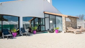 Ein modernes Gebäude mit großen Glasfenstern, einer Außenbereich-Sitzgelegenheit mit Stühlen und Topfpflanzen sowie einem Steinbau mit Ziegeldach.